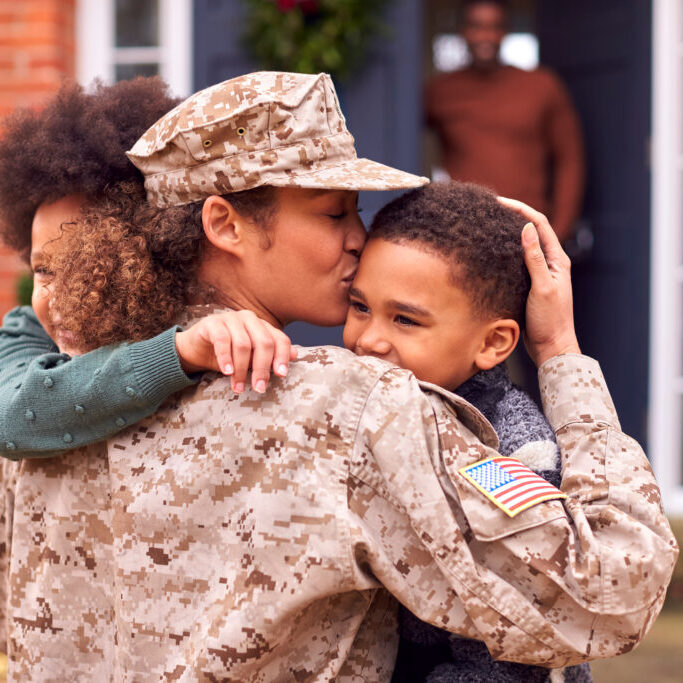 American Female Soldier In Uniform Returning Home To Family On Hugging Children Outside House