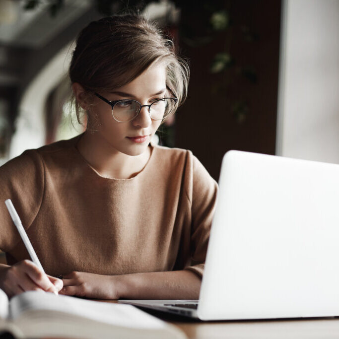 Creative good-looking european female with fair hair in trendy glasses, making notes while looking at laptop screen, working or preparing for business meeting, being focused and hardworking,